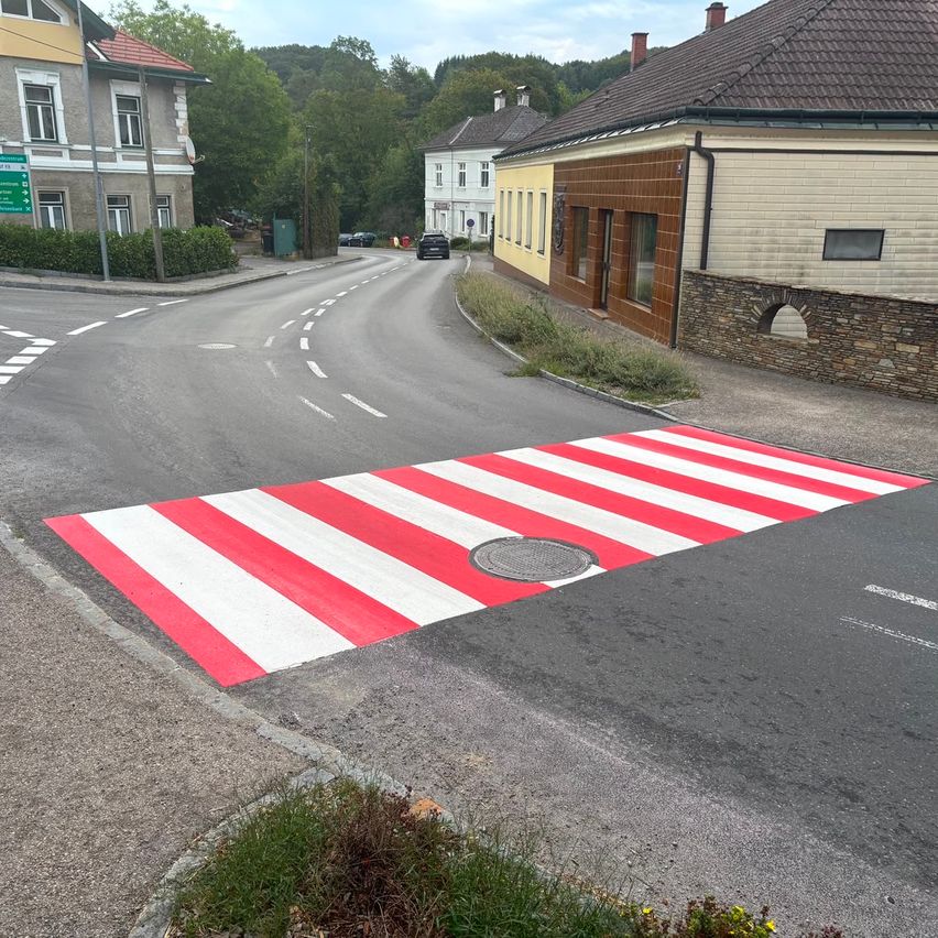 Fußgängerzone rot-weiß gestreift auf Asphalt mit Häusern und Bäumen im Hintergrund