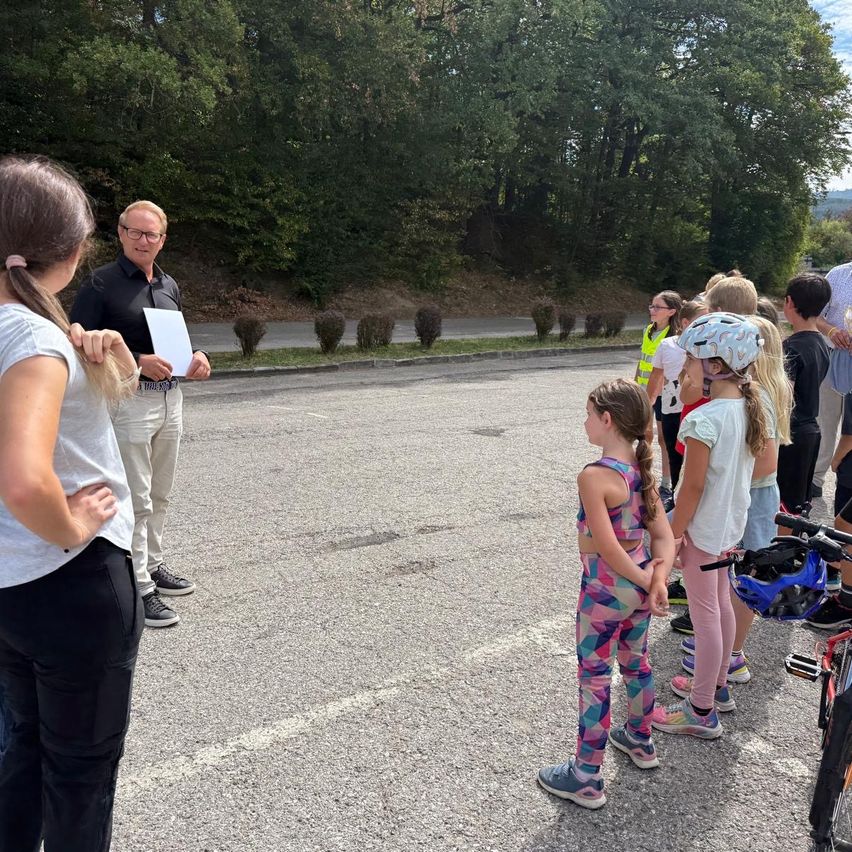 Ein Mann mit Brille und schwarzem Shirt spricht zu einer Gruppe von Kindern auf einem Parkplatz, einige mit Helmen.