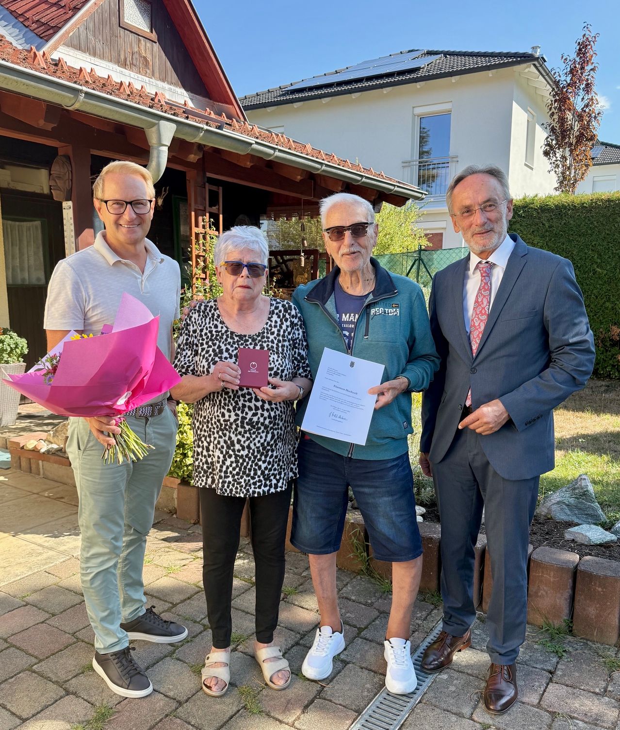 Vier Personen stehen vor einem Haus mit Garten. Der Mann links hält einen Blumenstrauß. Die Frau in der Mitte hält ein Zertifikat. Der Mann neben ihr trägt eine Sonnenbrille. Der Mann rechts trägt einen Anzug und eine Krawatte.