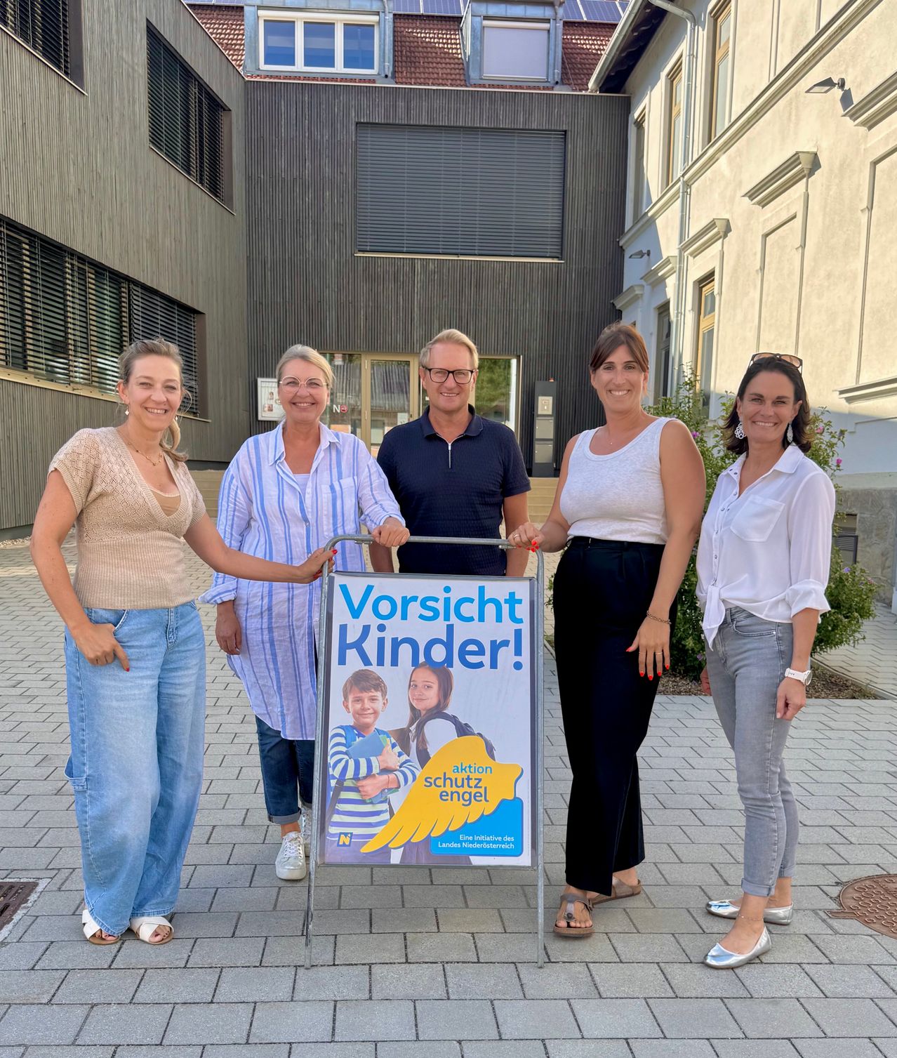 Fünf Personen stehen vor einem Gebäude mit einem Schild, das 'Vorsicht Kinder!' auf Deutsch besagt. Das Schild zeigt ein Bild eines Jungen und eines Mädchens.