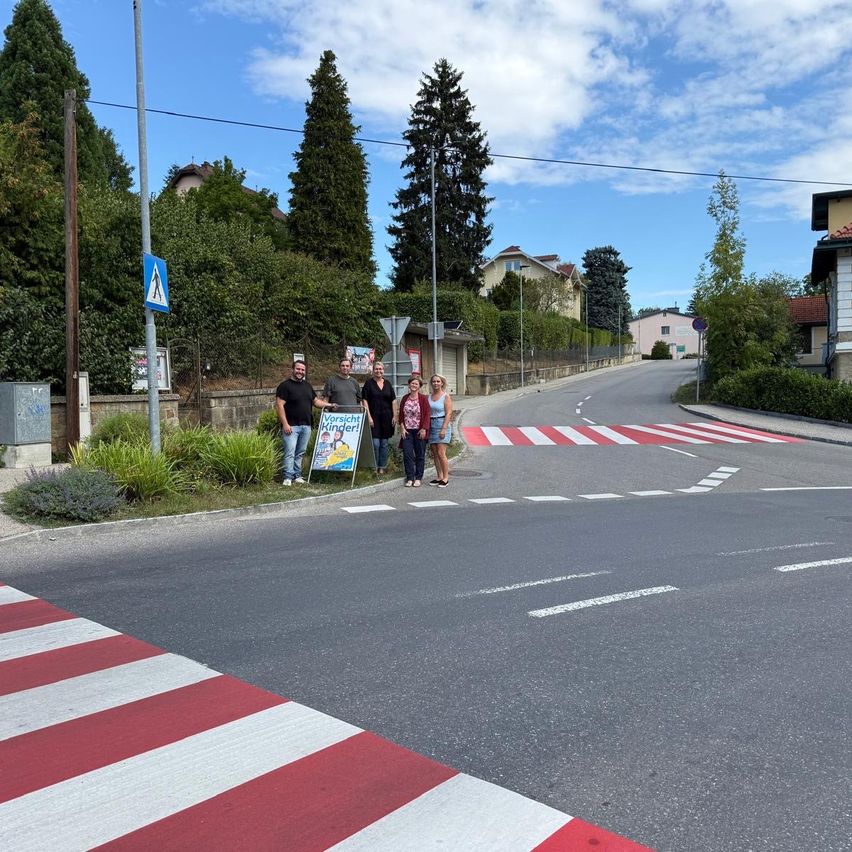 Fünf Personen stehen neben einem neu angelegten Zebrastreifen, der in Rot und Weiß auf einer Straße gemalt wurde.