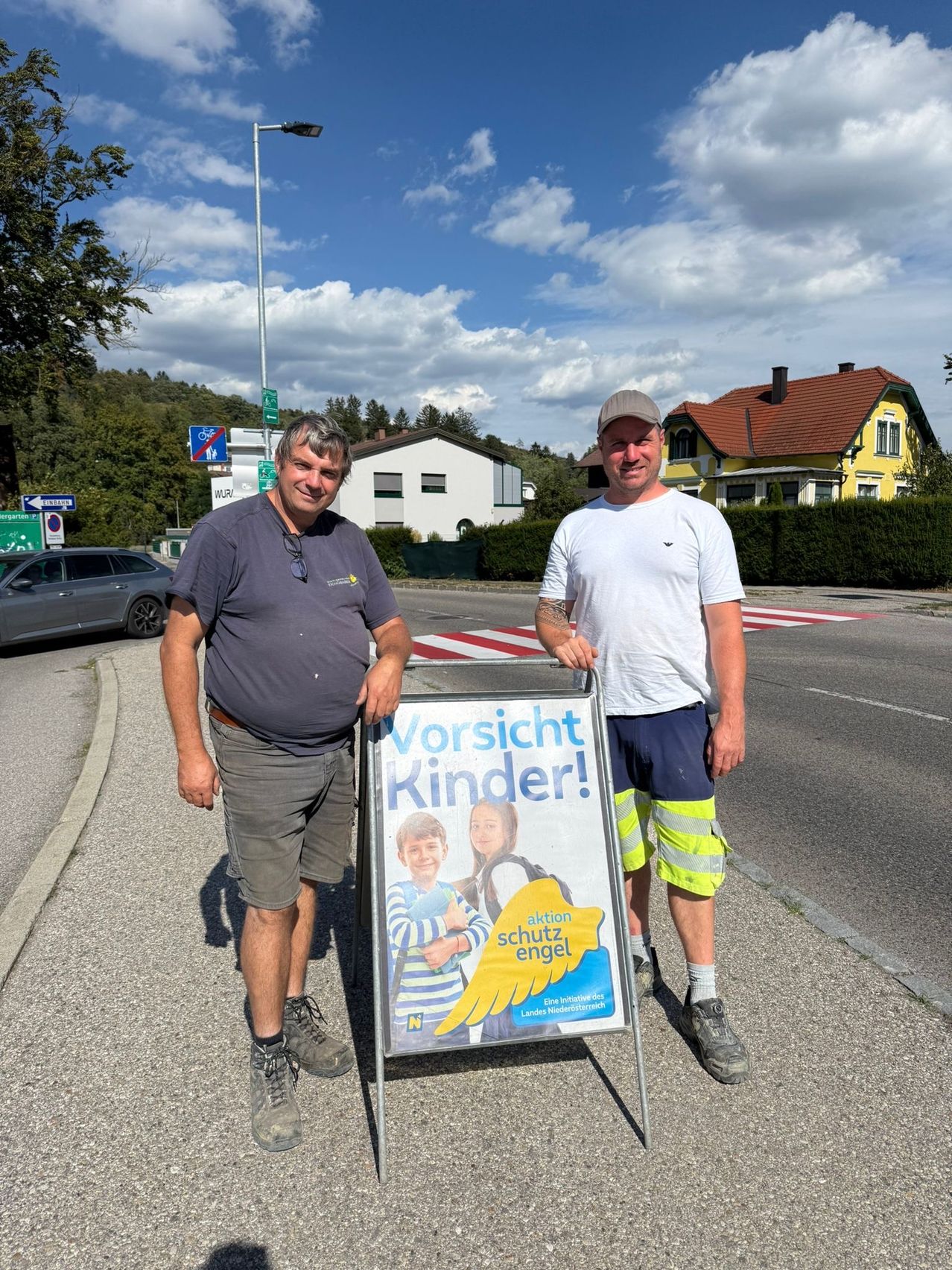 Zwei Männer stehen neben einem Schild, das 'Vorsicht Kinder!' in einem deutschsprachigen Land sagt. Ein Mann trägt einen Gürtel und Turnschuhe, der andere eine Mütze und reflektierende Shorts. Hinter ihnen sind ein Haus und ein Auto zu sehen.