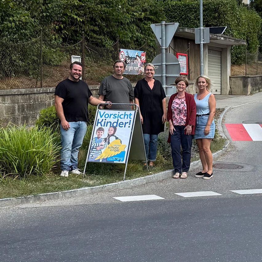 Fünf Personen stehen neben einem Schild, das Vorsicht Kinder! vor einer Straße steht.
