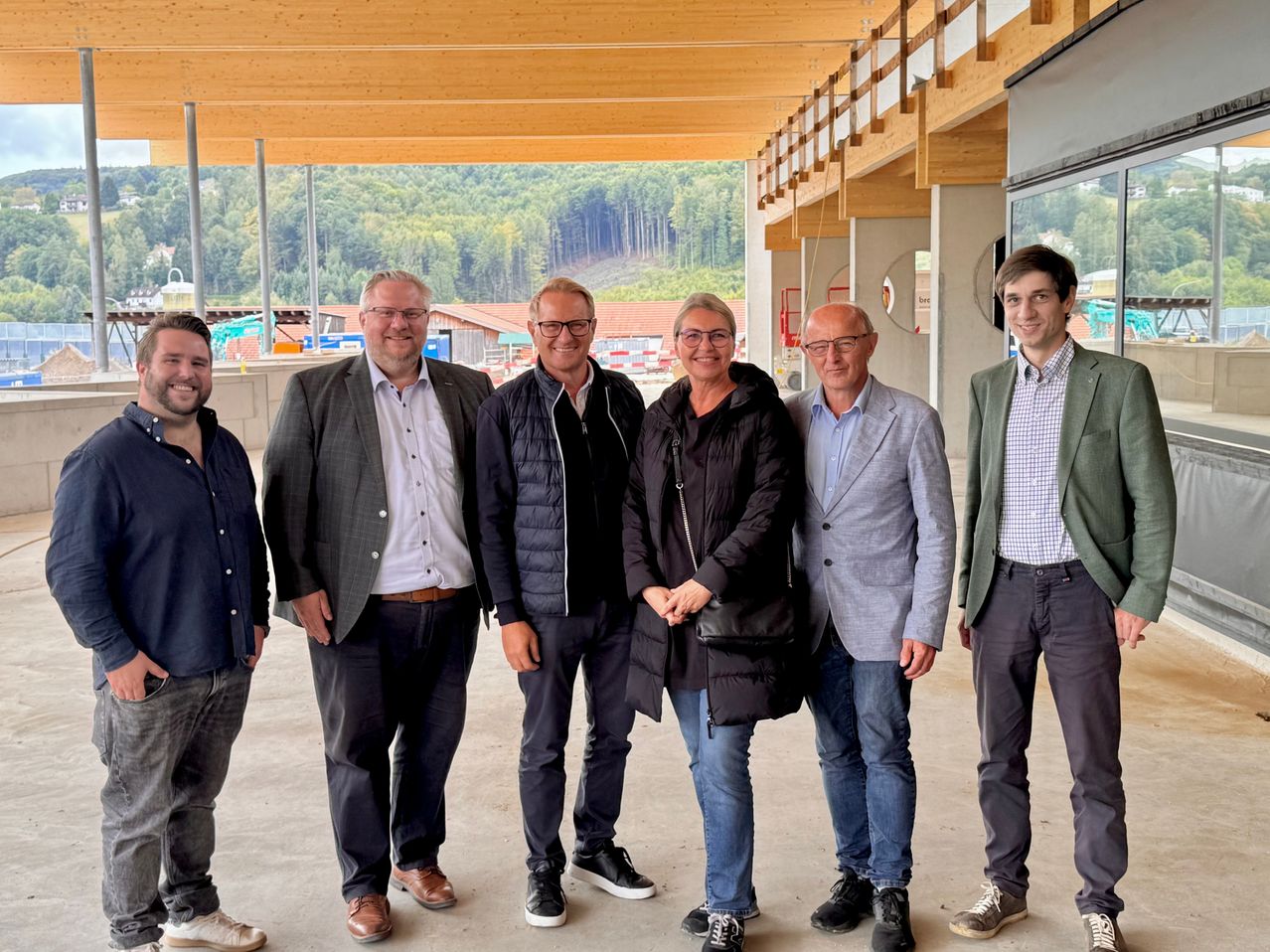 Sechs Personen, in unterschiedlicher Kleidung, stehen zusammen in einer Außenumgebung, möglicherweise für ein Gruppenfoto. Hinter ihnen ist eine Holzstruktur und eine Kulisse aus Bäumen und Bergen zu sehen.