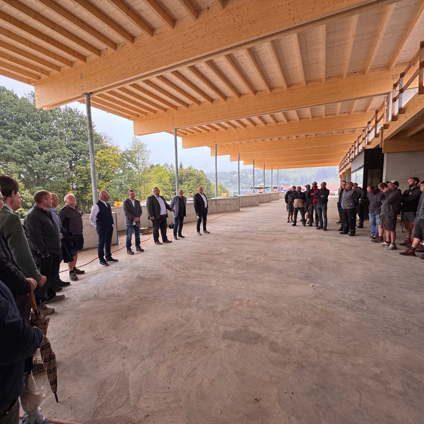Eine Gruppe von Menschen steht in einem großen, unfertigen Gebäude mit Holzbalken und Betonböden, unter einem klaren Himmel mit Bäumen im Hintergrund.