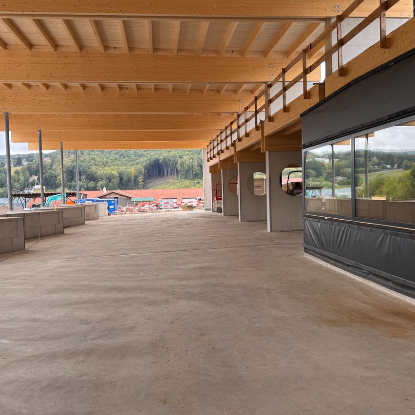 Ein leerer Bauplatz mit Holzbalken und Glasfenstern, mit Blick auf eine Landschaft mit Bäumen und einem Gebäude.