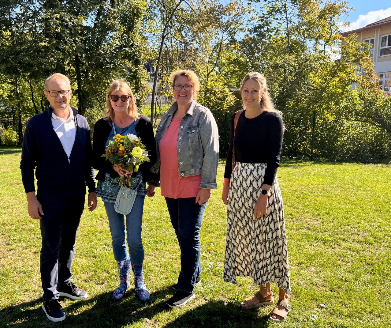 Vier Personen stehen zusammen auf einem Rasenfeld und lächeln für ein Foto. Die Frau auf der linken Seite hält einen Blumenstrauß. Hinter ihnen sind Bäume und ein Gebäude.