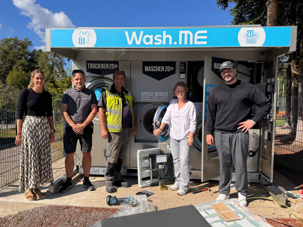 Fünf Personen stehen vor einer selbstbedienten Waschanlage mit der Aufschrift Wash.ME, zwei Männer in Warnwesten und zwei Männer und eine Frau in Freizeitkleidung.