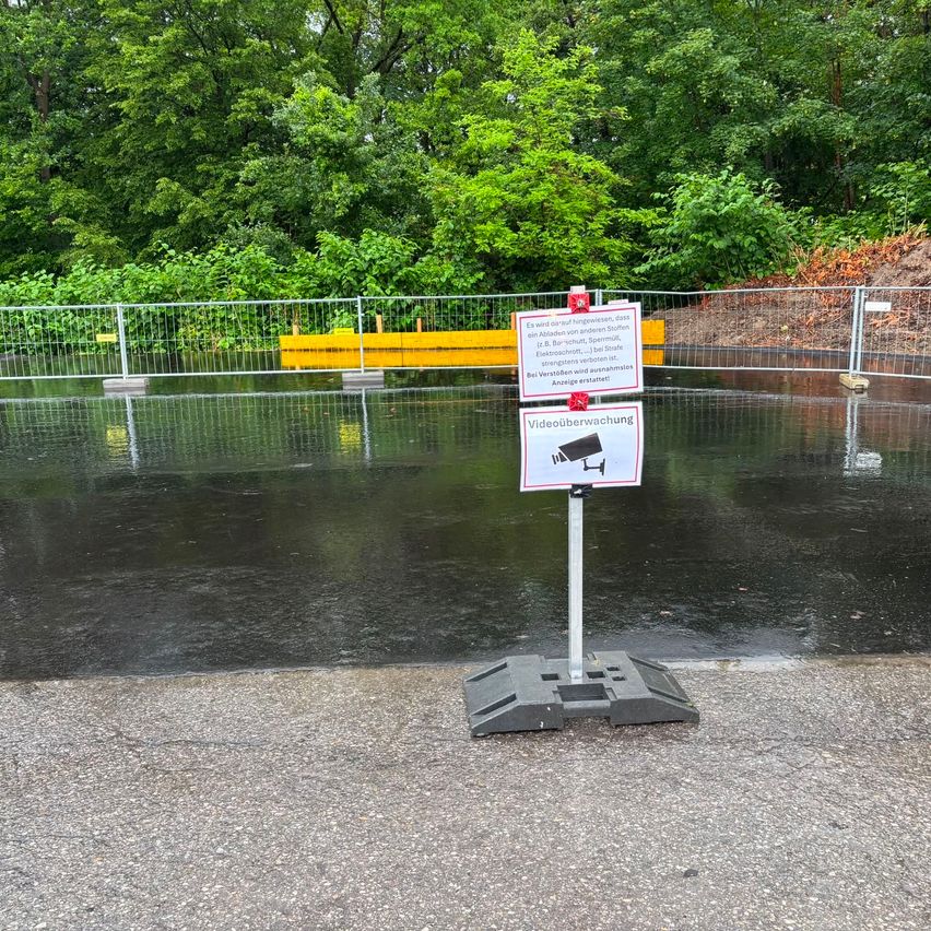 Ein nasser Bereich mit einem Warnschild für Videoüberwachung, umgeben von einem Metallzaun und Bäumen, mit Reflexionen auf dem Wasser.