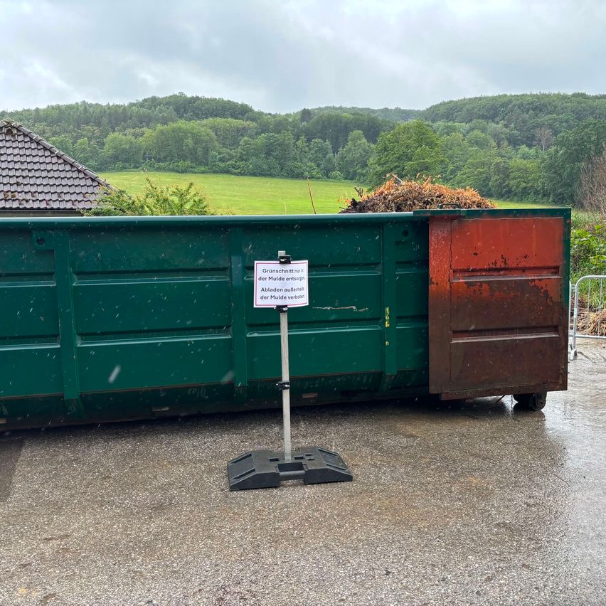 Ein großer grüner Container mit einem Schild, das 'Gruenschnitt fuer die Mulde entsorge der Mulde verbot' steht, befindet sich auf einer nassen Betonfläche mit einem Haus und Bäumen im Hintergrund.