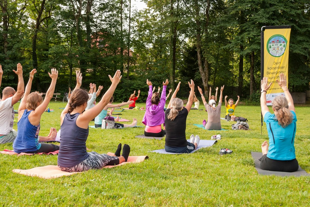 Eine Gruppe von Menschen praktiziert Yoga im Freien auf dem Rasen, umgeben von Bäumen. Einige tragen Freizeitkleidung, während andere Sportbekleidung tragen. Sie sitzen auf Yogamatten, und einige haben ihre Hände erhoben.