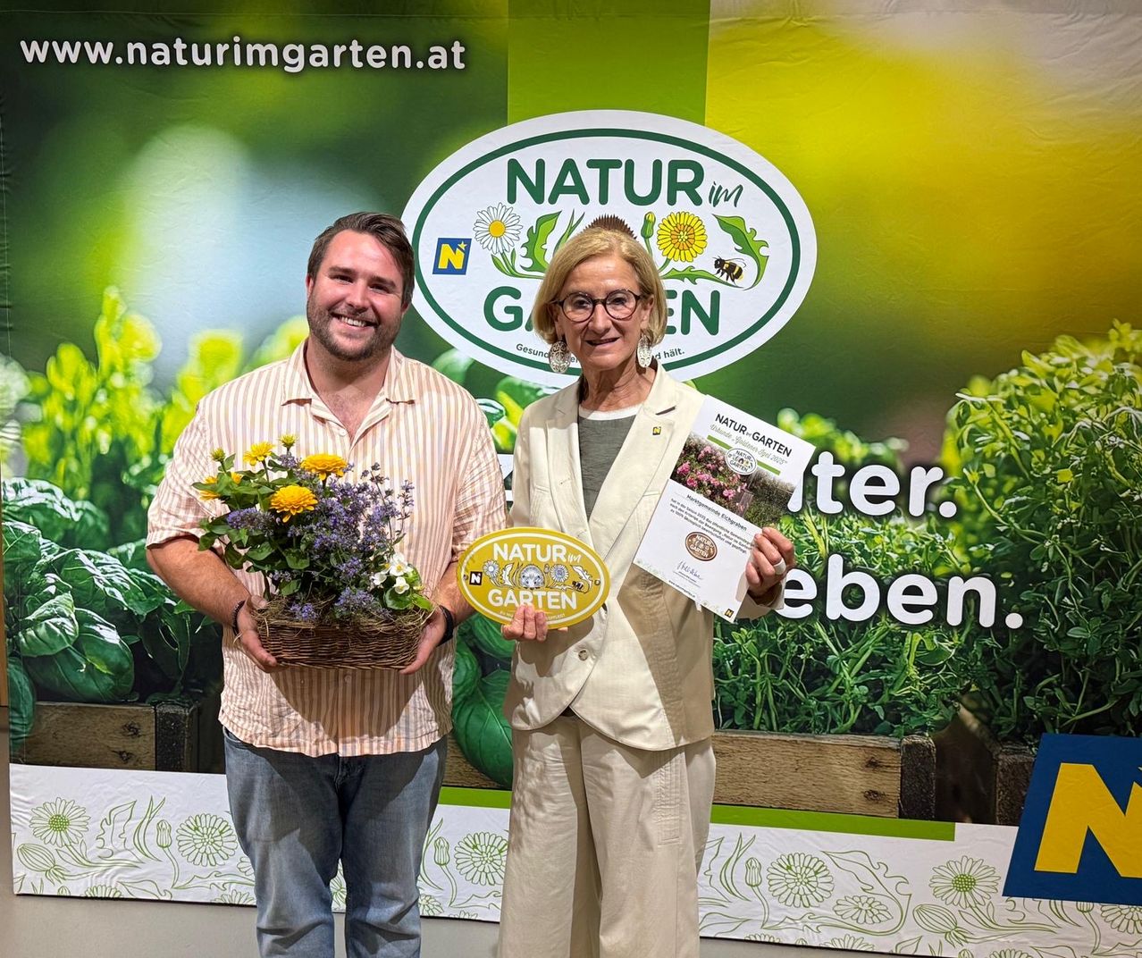 Ein Mann und eine Frau stehen vor einem grünen Banner mit den Worten 'Natur im Garten'. Der Mann hält einen Blumenstrauß, und die Frau hält ein Zertifikat.