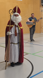 Ein Mann als Sankt Nikolaus verkleidet steht in einer Turnhalle. Ein Musiker spielt im Hintergrund die Gitarre.