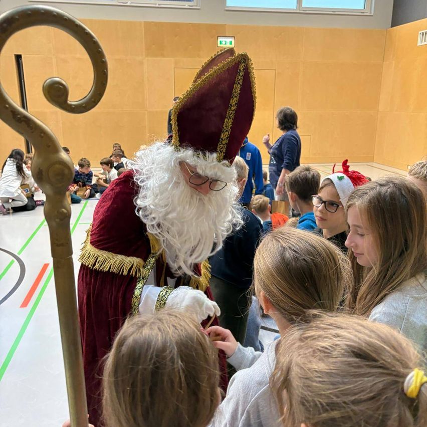 Eine Gruppe von Kindern ist um einen als Weihnachtsmann verkleideten Mann in einer Turnhalle versammelt. Die Kinder sehen ihn an, und er hält einen Stab. Hinter ihm ist eine Holzwand.