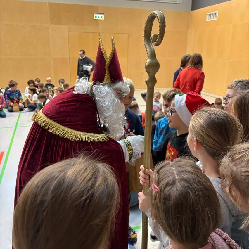 Ein Mann als Weihnachtsmann verkleidet hält einen Stab und interagiert mit Kindern in einem Raum.