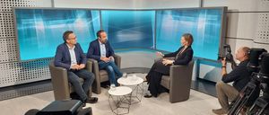 Drei Personen sitzen auf Stühlen in einem Studio. Die Frau gestikuliert während sie spricht. Zwei Männer sitzen neben ihr. Es gibt zwei kleine Tische mit Gläsern darauf.