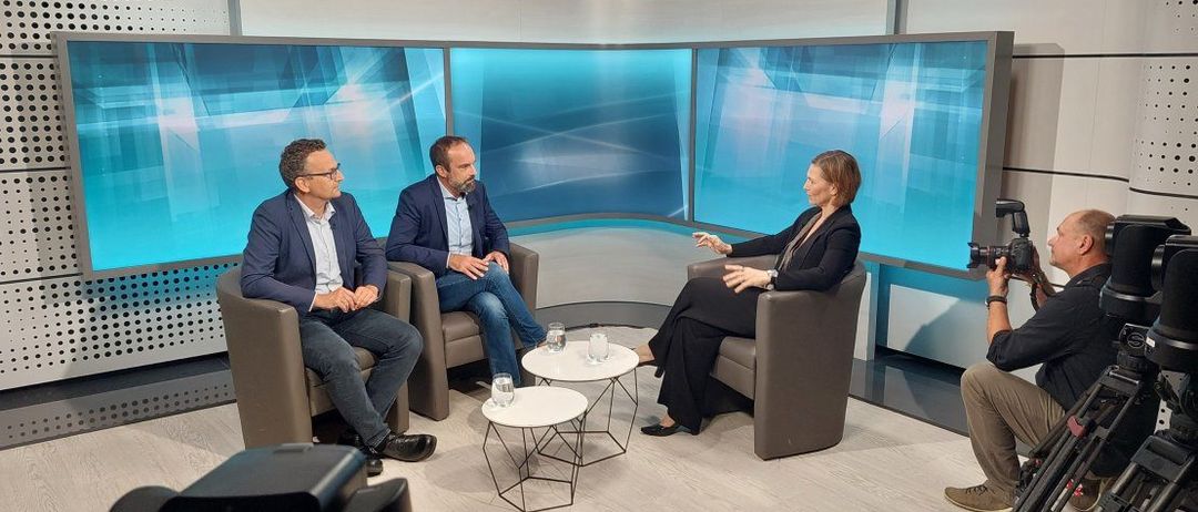 Drei Personen sitzen auf Stühlen in einem Studio. Die Frau gestikuliert während sie spricht. Zwei Männer sitzen neben ihr. Es gibt zwei kleine Tische mit Gläsern darauf.