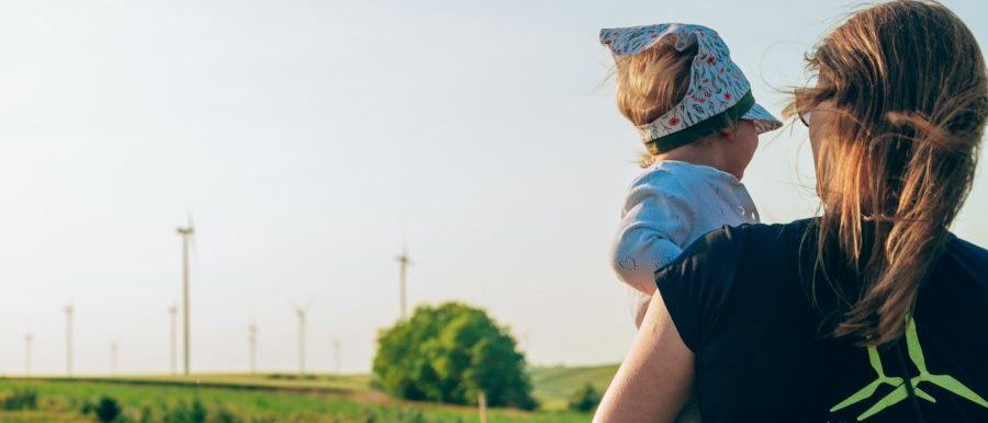 Eine Frau, die ein Kind hält, steht im Freien in der Nähe eines Feldes mit Bäumen und Windturbinen im Hintergrund.