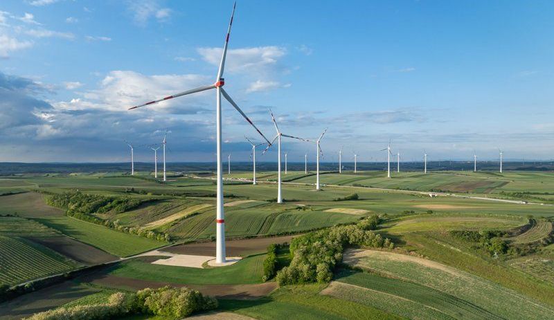 Luftaufnahme eines Windparks mit mehreren Windturbinen an einem sonnigen Tag, in einer grünen ländlichen Landschaft.