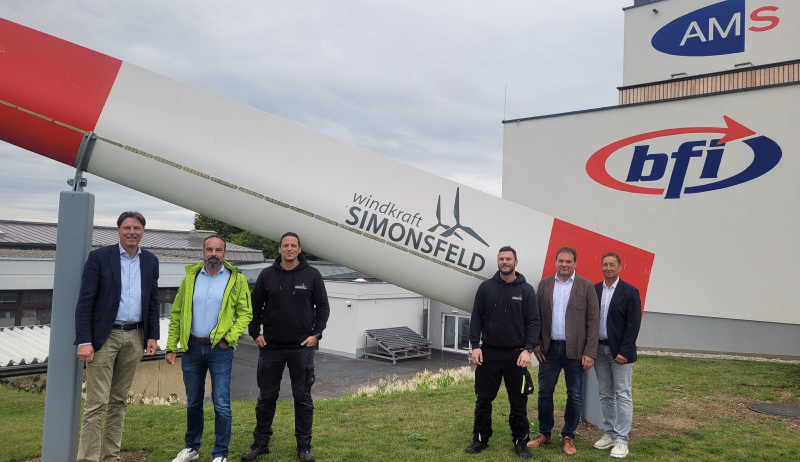 Fünf Männer stehen vor einer großen Windturbine, die mit den Worten 'windkraft SIMONSFELD' gekennzeichnet ist. Die Männer tragen Jacken und Hosen. Im Hintergrund befinden sich Gebäude mit Logos für AM und bfi.