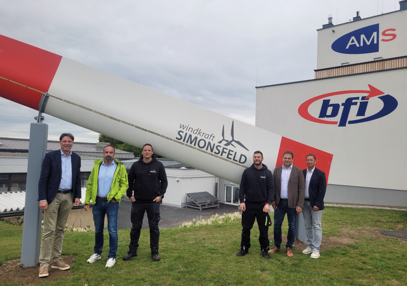 Fünf Männer stehen vor einer großen Windturbine, die mit den Worten 'windkraft SIMONSFELD' gekennzeichnet ist. Die Männer tragen Jacken und Hosen. Im Hintergrund befinden sich Gebäude mit Logos für AM und bfi.