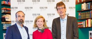 Drei Personen stehen gemeinsam vor einer Wand mit 'CEOs für die Zukunft' Text. Ein Mann in einem blauen Anzug und eine Frau in einem roten Kleid gehören dazu. Dahinter stehen Bücherregale voller Bücher.