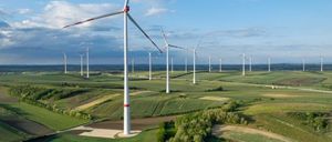 Eine Luftaufnahme eines weitläufigen Feldes mit mehreren Windturbinen, umgeben von grünen Pflanzen unter einem blauen Himmel mit verstreuten Wolken.