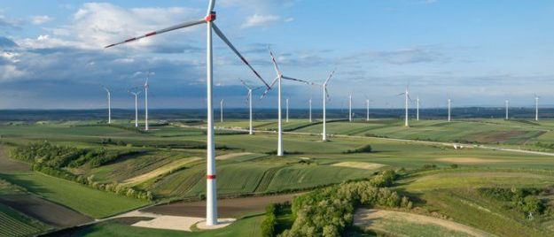 Eine Luftaufnahme eines weitläufigen Feldes mit mehreren Windturbinen, umgeben von grünen Pflanzen unter einem blauen Himmel mit verstreuten Wolken.