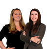 Zwei Frauen stehen eng beieinander und lächeln für ein Bild. Die Frau auf der linken Seite hat lange blonde Haare und trägt ein schwarzes Oberteil. Die Frau auf der rechten Seite hat braune Haare und trägt einen grauen Blazer. Beide sind professionell gekleidet.