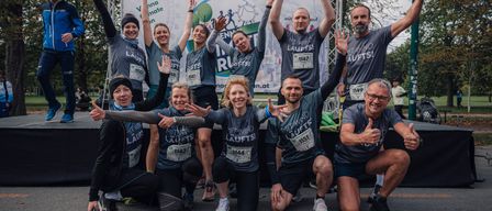 Eine Gruppe von Läufern, sowohl Männer als auch Frauen, posiert für ein Foto mit erhobenen Händen und Lächeln, tragen passende graue T-Shirts mit dem Text 'Mit Wind Laufts' und Nummern auf ihren Trikots.