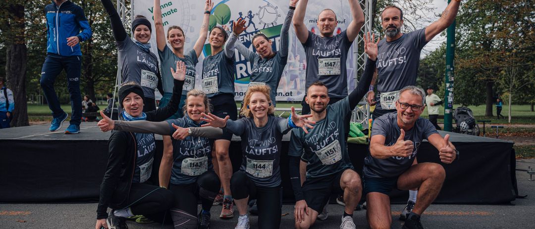 Eine Gruppe von Läufern, sowohl Männer als auch Frauen, posiert für ein Foto mit erhobenen Händen und Lächeln, tragen passende graue T-Shirts mit dem Text 'Mit Wind Laufts' und Nummern auf ihren Trikots.