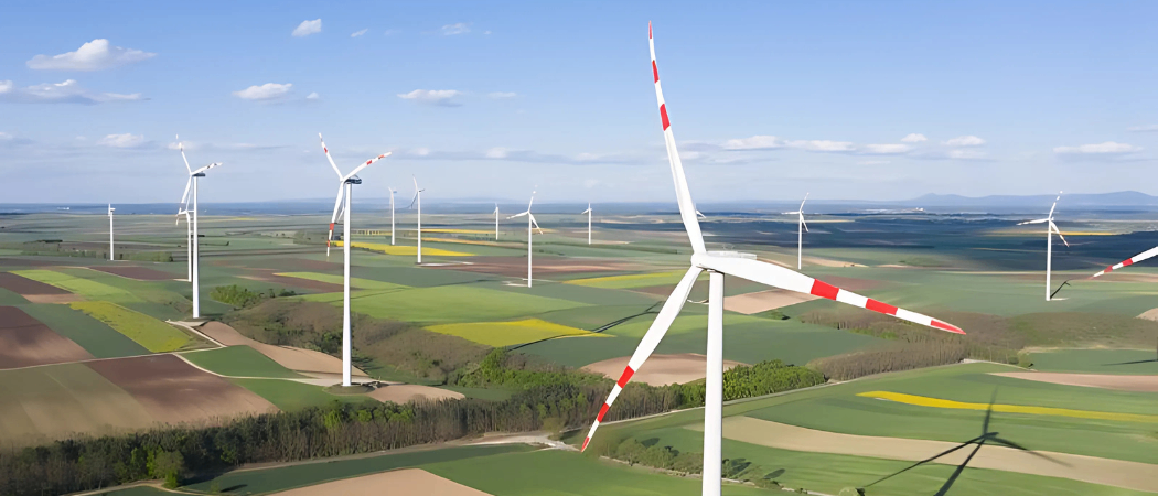 Luftaufnahme eines Feldes mit mehreren Windturbinen unter einem klaren Himmel. Die Turbinen haben rote und weiße Rotorblätter. Das Feld ist in Abschnitte mit grünen und gelben Pflanzen unterteilt.