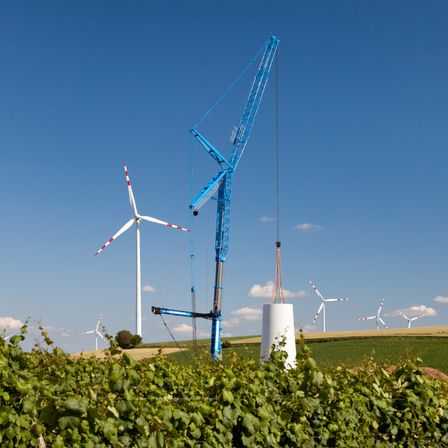 Ein blauer Kran hebt einen großen zylindrischen Gegenstand in der Nähe eines Windrads. Im Hintergrund sind mehrere weitere Windräder zu sehen.