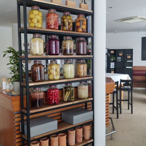 Ein hoher Regalstand voller verschiedener Gläser mit eingelegtem Obst und Gemüse, eine Pflanze links und ein Tisch mit einem Stuhl rechts.