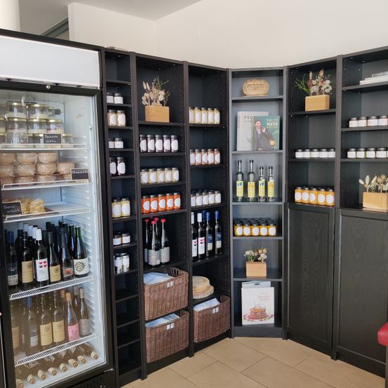 Ein kleiner Lebensmittelladen mit einem offenen Kühlschrank, der Flaschen Wein und Käse zeigt. Regale mit verschiedenen Gläsern und Flaschen, und dekorative Pflanzen schmücken die Ecken.