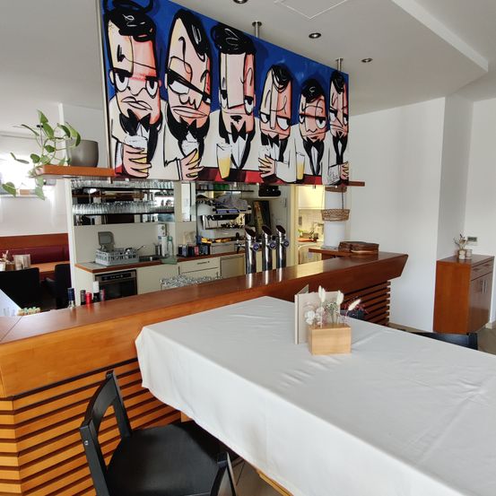 Eine Bar mit einem großen Wandbild von Cartoon-Männern darüber. Eine weiße Tischdecke ist auf dem Tresen der Bar mit einer kleinen Holzbox mit Blumen gedeckt.