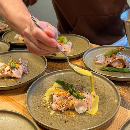 Ein Koch bereitet ein Gericht mit Lachs, Spargel und gelber Sauce zu. Er benutzt einen Löffel. Der Tisch hat mehrere Teller mit ähnlichen Gerichten.