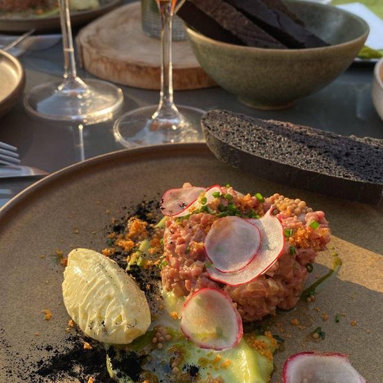 Ein Teller mit Thunfisch-Tartare, garniert mit Radieschenscheiben und grünen Zwiebeln, neben einer Zitronenscheibe, serviert mit Brot in einer Schüssel und Weingläsern.