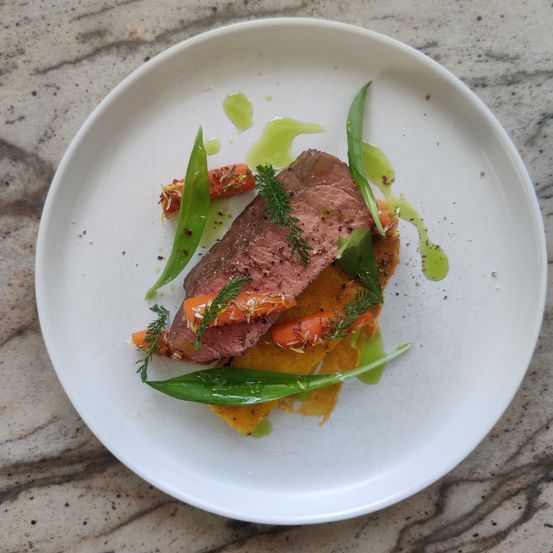 Ein weißer Teller präsentiert ein Gourmetgericht mit einem Stück Fleisch, das mit Kräutern und grünen Bohnen garniert ist, zusammen mit einer Sauce und einer Karotte. Das Gericht wird auf einer Marmoroberfläche serviert.