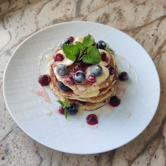 Ein weißer Teller enthält einen Stapel Pfannkuchen, überzogen mit Blaubeeren, Himbeeren, Bananen und Minzblättern, mit karamellisiertem Sirup auf der Seite.