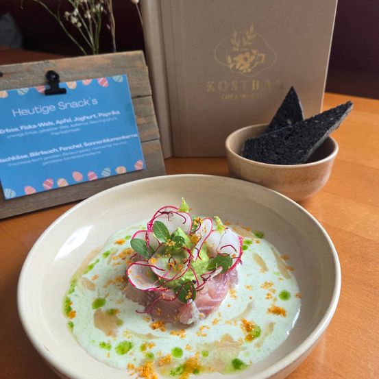Ein Gericht mit Thunfisch, Radieschen und Avocado steht auf einem Holztisch. Ein Schild steht Heuteige Snack's. Dahinter befindet sich eine Box mit der Aufschrift Kostbar Cafe & Bar.