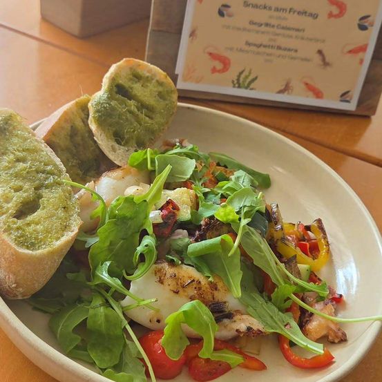 Ein Teller mit gegrilltem Fisch, Gemüse und Rucolasalat, begleitet von zwei Scheiben Pesto-betopftem Brot, steht auf einem Holztisch.