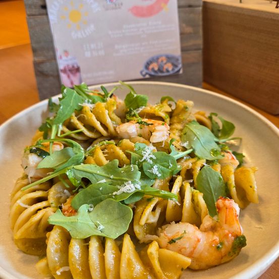 Eine Platte mit Spiralnudeln, überzogen mit Garnelen, Rucola und Käse. Im Hintergrund befindet sich ein Holzschild mit Logo und Text.