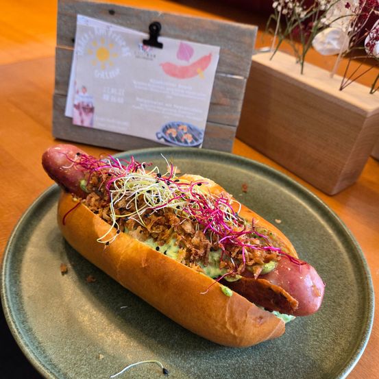 Ein Hot Dog mit Avocado, Sprossen und knusprigen Zwiebeln wird auf einem grünen Teller serviert. Ein Holzschild und eine Holzbox mit Blumen sind im Hintergrund.