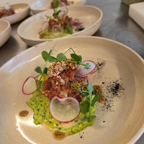Ein Gericht mit einem Fleischpatty, garniert mit Radieschen, Kräutern und grüner Sauce, präsentiert auf einem weißen Teller mit zusätzlichen Gerichten im Hintergrund.