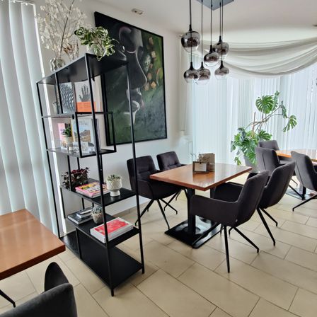 Ein moderner, offener Essbereich mit einem Holztisch, Stühlen und einem schwarzen Regal mit Büchern und Pflanzen. Der Raum hat weiße Wände, Pendelleuchten und ein großes Gemälde.