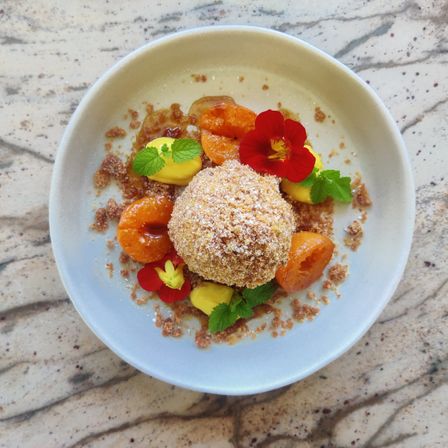 Eine weiße Schüssel mit einem Dessert, bestehend aus einer knusprigen Kugel, Orangensegmenten, roten Blumen und Minzblättern auf einer Marmoroberfläche.