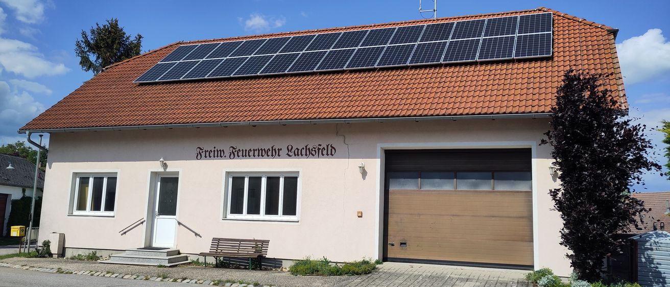 Ein rosafarbenes Gebäude mit rotem Dach und Solarpanelen hat eine Bank, eine Garage und ein Schild mit der Aufschrift Freiwillige Feuerwehr Lachsfeld.