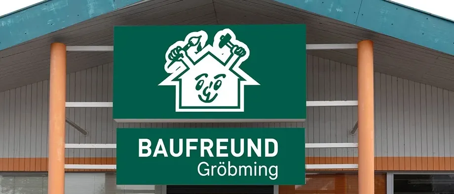 Die Vorderseite eines Gebäudes hat ein Schild mit der Aufschrift Baufreund Grobming. Das Schild ist grün mit weißen Buchstaben. Das Gebäude hat eine Glastür und Fenster.