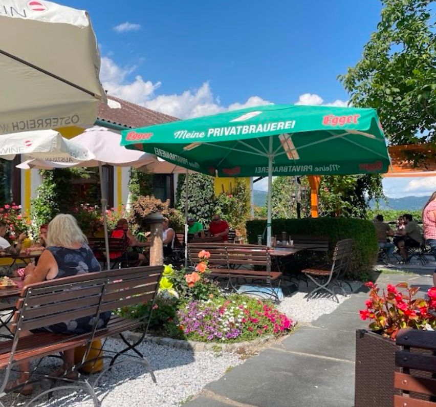 Eine Frau sitzt auf einer Bank in einem Restaurantgarten mit bunten Blumen, unter einem grünen Schirm. Das Restaurant hat Holzbänke und Tische.